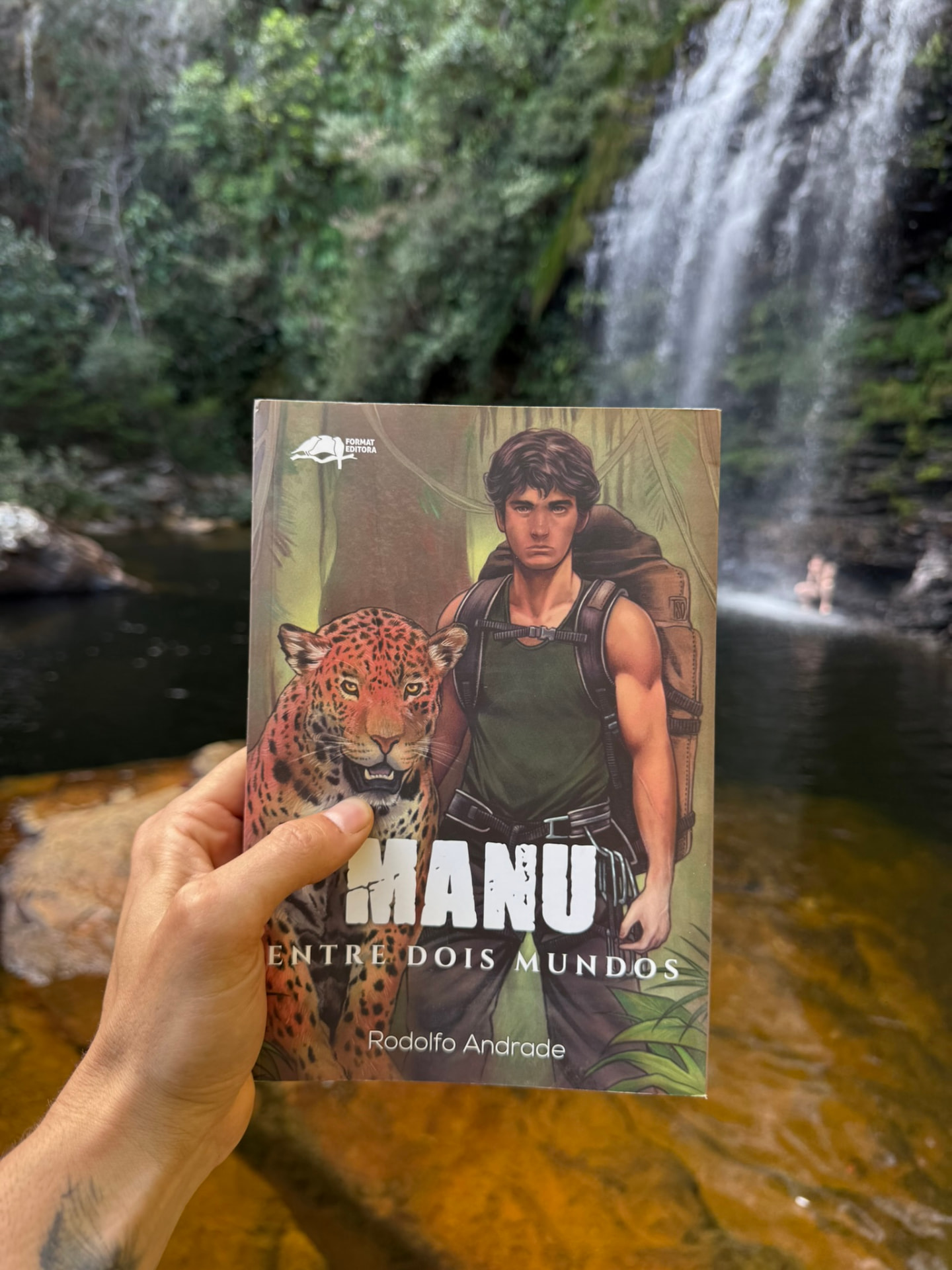 Livro Manu segurado em frente a uma cachoeira
